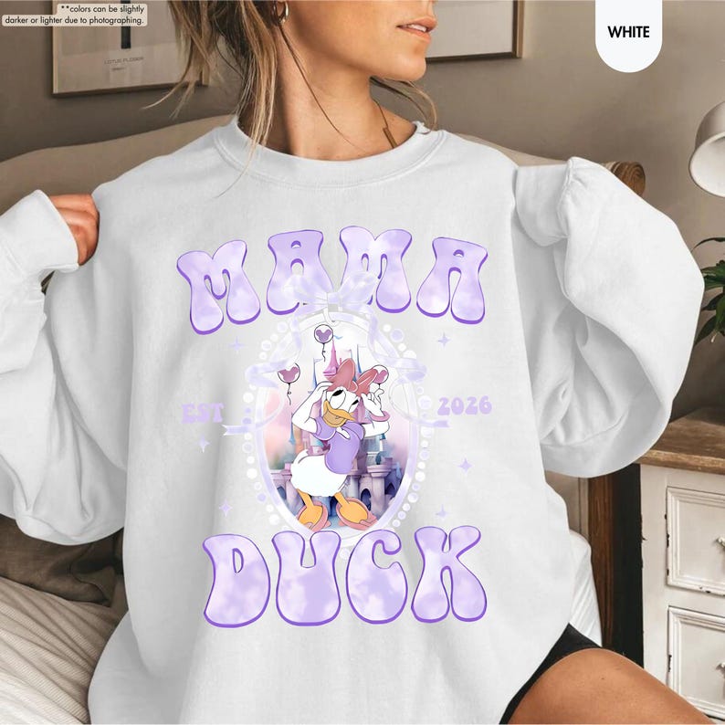 May include: White sweatshirt with the text "MAMA DUCK" in purple bubble letters. The design includes a cartoon image of Daisy Duck, a castle, and the year 2026. The sweatshirt is a casual, comfortable style.