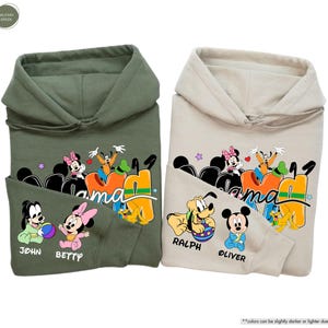 K&ouml;nnte beinhalten: Zwei Kapuzenpullover, einer in Milit&auml;rgr&uuml;n und einer in Sandfarbe, mit einer bunten Grafik von Disney-Figuren. Auf den Sweatshirts sind die W&ouml;rter "Mama" und Namen wie "John", "Betty", "Ralph" und "Oliver" aufgedruckt.