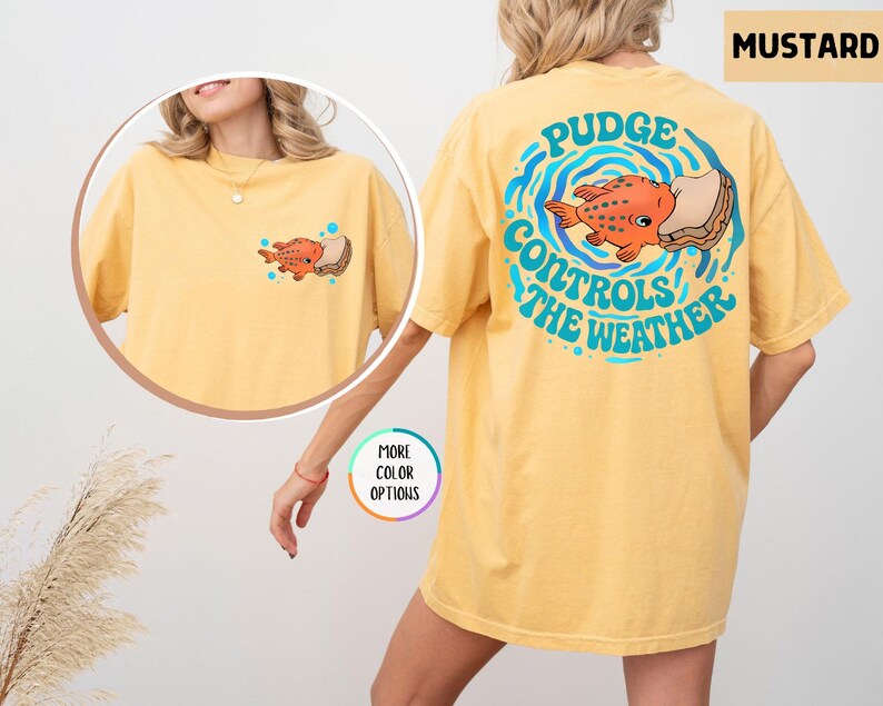 Puede incluir: Camiseta de color mostaza con un pez de dibujos animados y el texto "Pudge Controls the Weather" en la espalda. La parte delantera de la camiseta tiene un gr&aacute;fico de pez m&aacute;s peque&ntilde;o. La camiseta tiene un corte holgado.