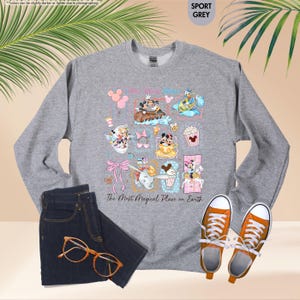 May include: Sport grey sweatshirt featuring colorful Disney character illustrations. The design includes Mickey and Minnie Mouse, Donald Duck, and other characters. The text "The Most Magical Place on Earth" is printed below the illustrations. Paired with blue jeans and orange sneakers.