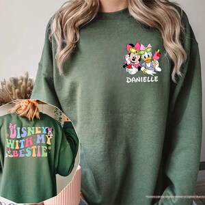 May include: A green sweatshirt with a colorful graphic of Disney characters Mickey Mouse and Minnie Mouse. The text "DISNEY WITH MY BESTIE" is printed on the back of the sweatshirt. The text "DANIELLE" is printed on the front of the sweatshirt.