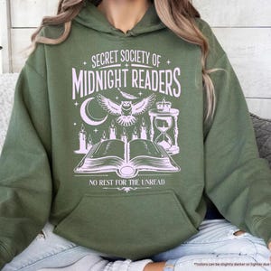 May include: Military green hoodie with a front graphic that reads "SECRET SOCIETY OF MIDNIGHT READERS" with an owl, moon, candles, and an open book. The text "NO REST FOR THE UNREAD" is below the graphic. The hoodie has a kangaroo pocket.