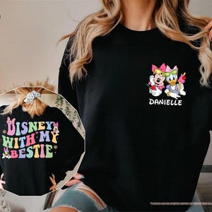 May include: Black sweatshirt with a colorful graphic of Disney characters Minnie Mouse and Daisy Duck wearing sunglasses and holding drinks. The text "Disney with my bestie" is on the back of the sweatshirt. The name "Danielle" is printed below the characters.