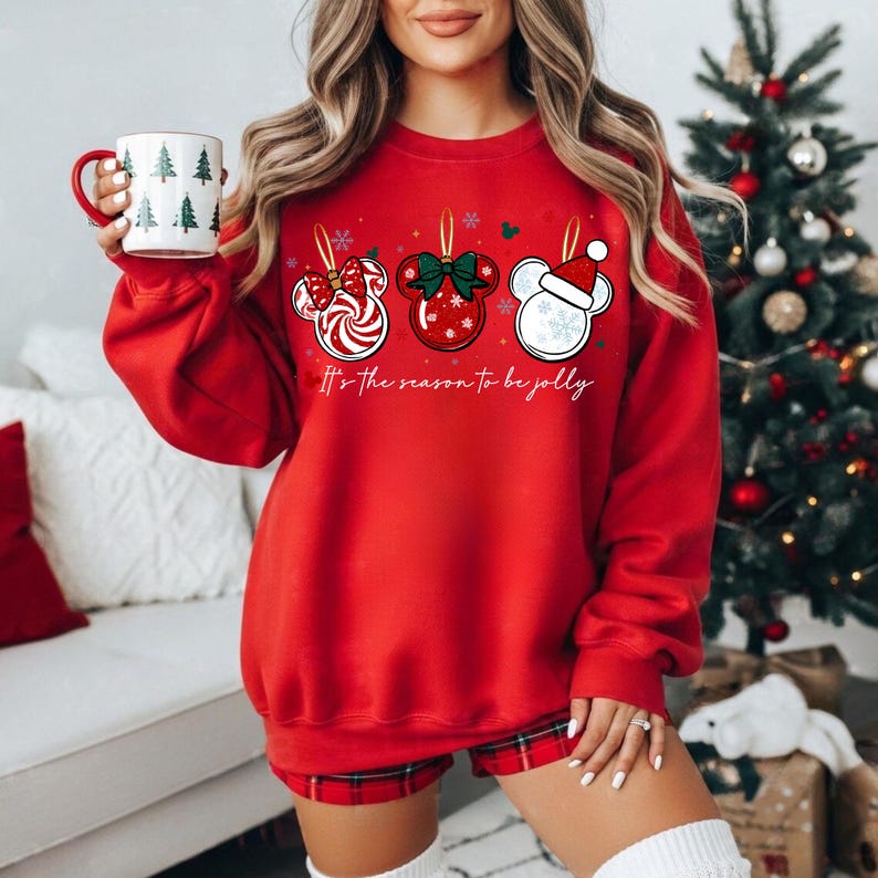 May include: Red sweatshirt with three Mickey Mouse-shaped Christmas ornament designs. The ornaments feature a candy cane, a red bow, and a Santa hat. The text "It's the season to be jolly" is printed below the ornaments. A white mug with Christmas trees is held in the hand.