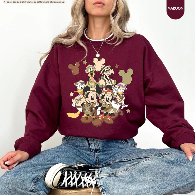 May include: Maroon sweatshirt featuring a graphic of Disney characters in safari attire. The design includes Mickey and Minnie Mouse, Donald Duck, Goofy, and Pluto. The sweatshirt has a crew neck and long sleeves. The word "MAROON" is printed on the upper right corner.