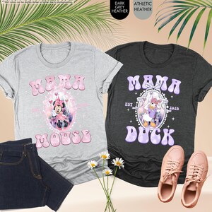 May include: Two graphic t-shirts, one gray and one dark gray, with the words "Mama Mouse" and "Mama Duck" in bubble letters. The gray shirt features Minnie Mouse, and the dark gray shirt features Daisy Duck. A pair of jeans and sneakers are also in the image.