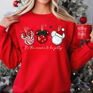 May include: A red sweatshirt featuring three Mickey Mouse-shaped Christmas ornaments with festive designs. The ornaments include a candy cane bow, a green bow, and a Santa hat. The text "It's the season to be jolly" is printed below the ornaments.