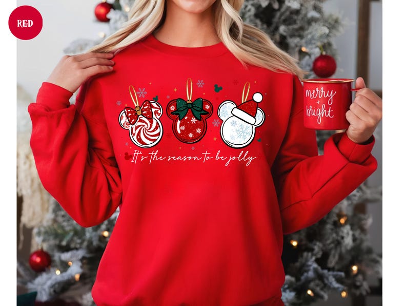 May include: A red sweatshirt featuring three Mickey Mouse-shaped Christmas ornaments with festive designs. The ornaments include a candy cane bow, a green bow, and a Santa hat. The text "It's the season to be jolly" is printed below the ornaments.
