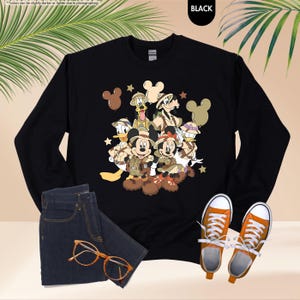 May include: Black long-sleeve sweatshirt featuring a colorful graphic of Disney characters in safari attire. The design includes Mickey and Minnie Mouse, Donald Duck, Goofy, and others. Paired with blue jeans, orange-laced sneakers, and orange-framed glasses.