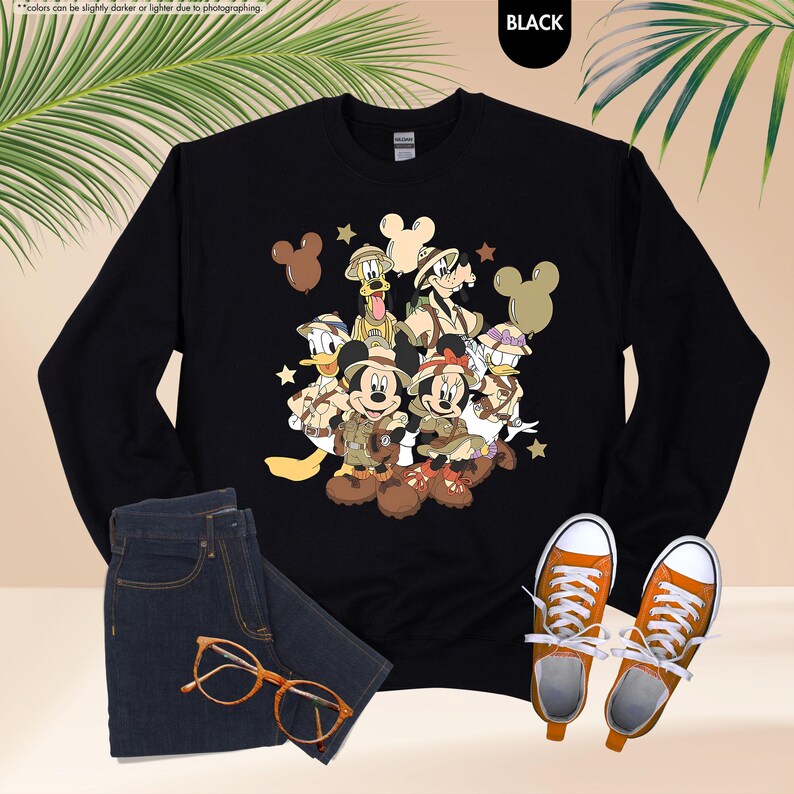 May include: Black long-sleeve sweatshirt featuring a colorful graphic of Disney characters in safari attire. The design includes Mickey and Minnie Mouse, Donald Duck, Goofy, and others. Paired with blue jeans, orange-laced sneakers, and orange-framed glasses.