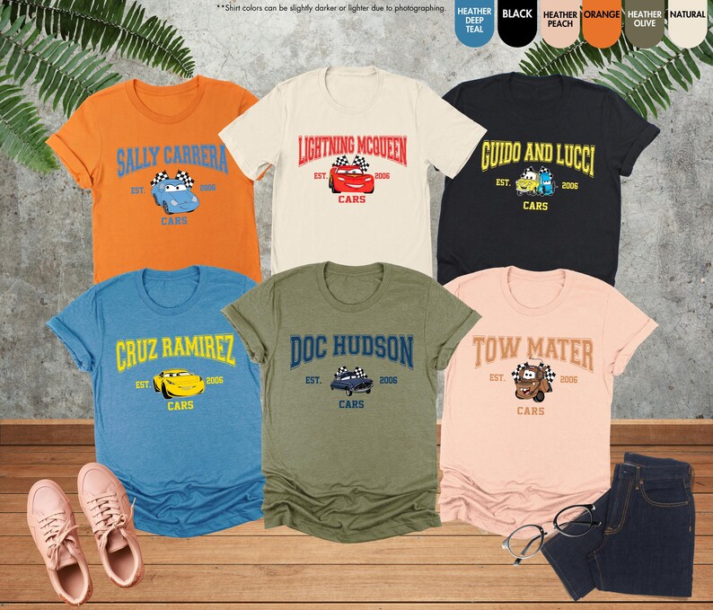 Puede incluir: Una colecci&oacute;n de camisetas con tem&aacute;tica de Cars en varios colores, incluyendo naranja, crema, negro, azul, verde oliva y melocot&oacute;n. Cada camiseta presenta el nombre y la imagen de un personaje, junto con el texto "Cars" y el a&ntilde;o "2006".