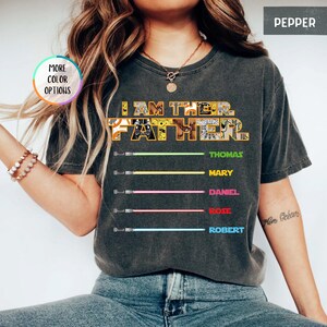 Puede incluir: Camiseta gris oscuro con el texto "I AM THE FATHER" en un dise&ntilde;o amarillo y blanco. Debajo del texto hay cinco sables de luz de colores con los nombres: Thomas, Mary, Daniel, Rose y Robert.