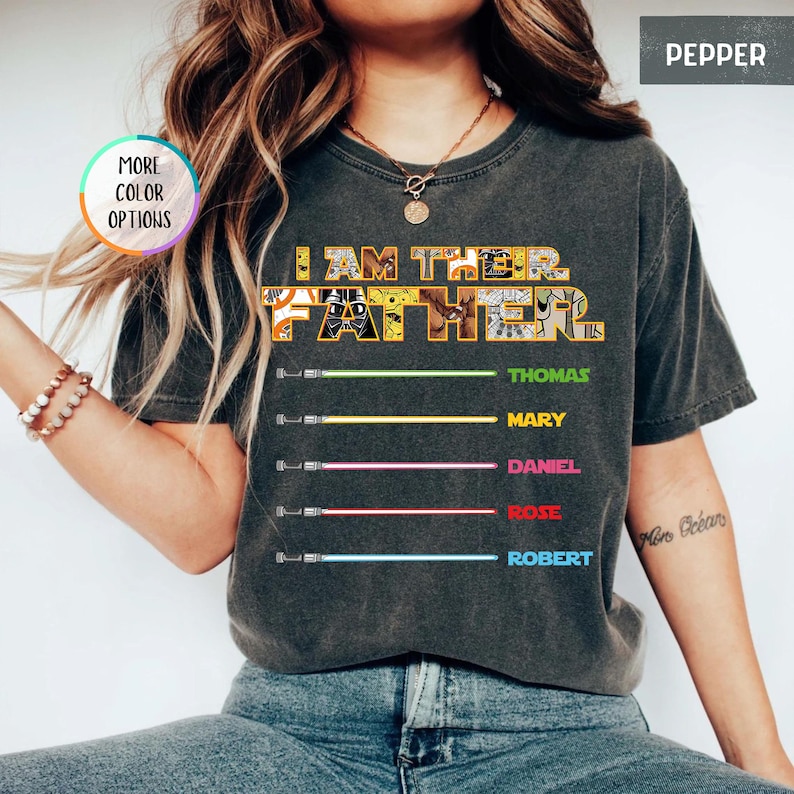 Puede incluir: Camiseta gris oscuro con el texto "I AM THE FATHER" en un dise&ntilde;o amarillo y blanco. Debajo del texto hay cinco sables de luz de colores con los nombres: Thomas, Mary, Daniel, Rose y Robert.
