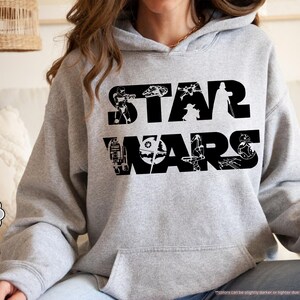 May include: A light grey hoodie with the words "STAR WARS" in large black letters. The letters are filled with black silhouettes of characters and vehicles from the Star Wars franchise. The hoodie has a front pocket and a hood.