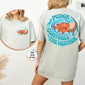 Puede incluir: Camiseta gris claro con un dise&ntilde;o de pez de dibujos animados. La parte posterior de la camiseta tiene un gr&aacute;fico m&aacute;s grande con el texto "Pudge Controls The Weather" en azul y blanco. La parte delantera tiene una versi&oacute;n m&aacute;s peque&ntilde;a del dise&ntilde;o del pez.