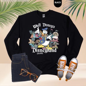 May include: Black sweatshirt featuring a colorful graphic of Donald Duck in front of Disneyland landmarks. The text "Walt Disney's Disneyland Est.1955" is printed below the graphic. The image also includes a pair of blue jeans, orange glasses, and orange sneakers.