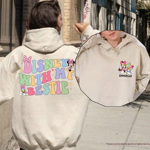 May include: Two cream colored hoodies with Disney character graphics. One hoodie says "Disney with my bestie" and features Minnie Mouse and Daisy Duck. The other hoodie says "Danielle" and features Minnie Mouse, Daisy Duck, and Donald Duck.