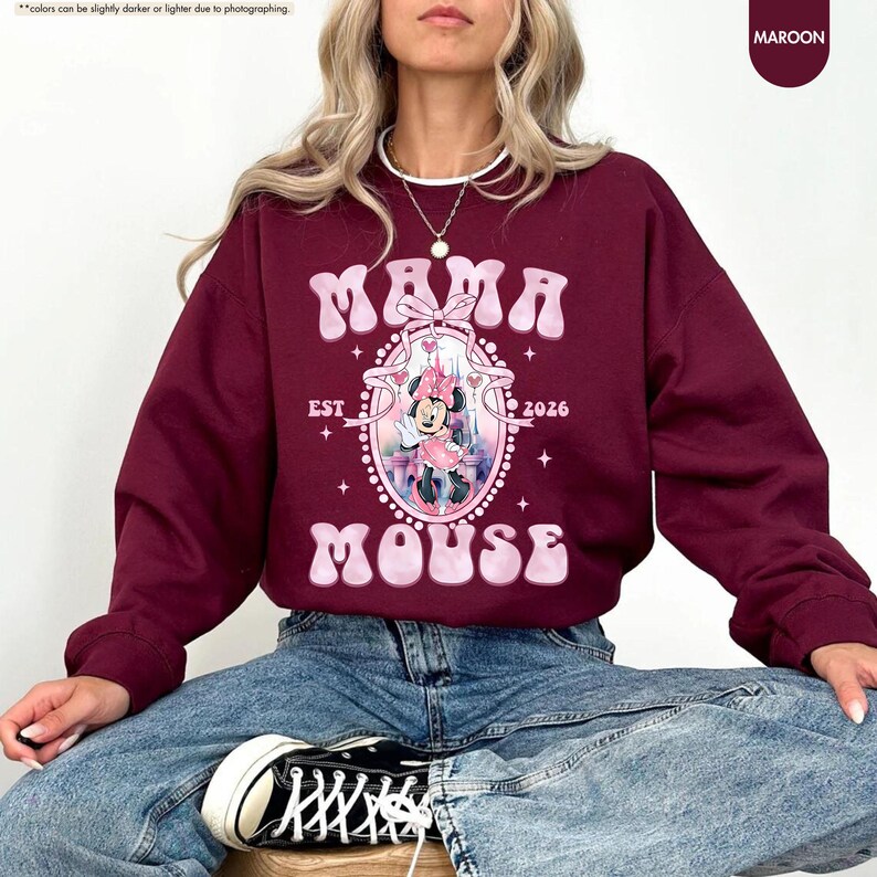 May include: Maroon sweatshirt with the text "MAMA MOUSE EST 2026" in a retro font. The graphic features a cartoon mouse in a pink dress. The sweatshirt is a solid maroon color.
