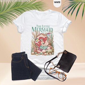 May include: White t-shirt featuring "The Little Mermaid" graphic. The design includes Ariel, Flounder, and other sea creatures. The shirt is paired with dark blue jeans, a black purse, and brown eyeglasses.