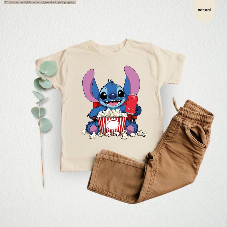 May include: A beige t-shirt featuring a cartoon character, holding a red drink and a popcorn bucket. The character is blue with large ears. Brown pants are next to the shirt. The shirt and pants are on a white background.