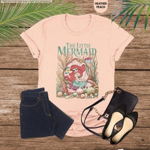 May include: Heather peach t-shirt featuring "The Little Mermaid" graphic. The design includes Ariel, Flounder, and other sea creatures. Paired with dark wash jeans, black purse, and black flats.