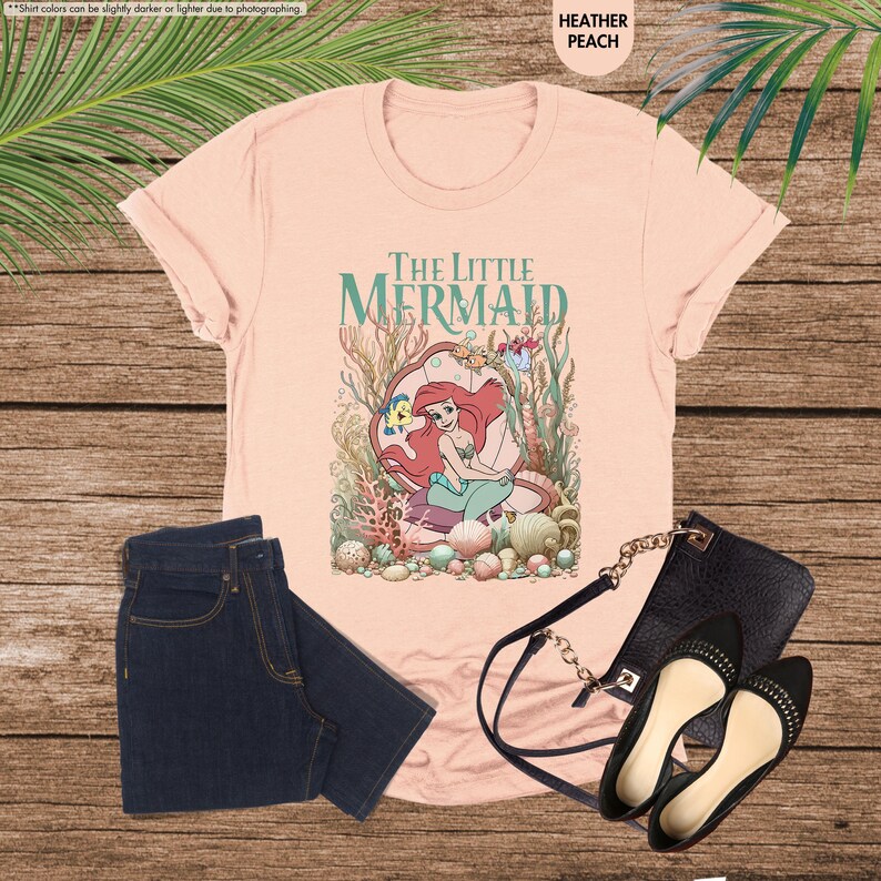 May include: Heather peach t-shirt featuring "The Little Mermaid" graphic. The design includes Ariel, Flounder, and other sea creatures. Paired with dark wash jeans, black purse, and black flats.
