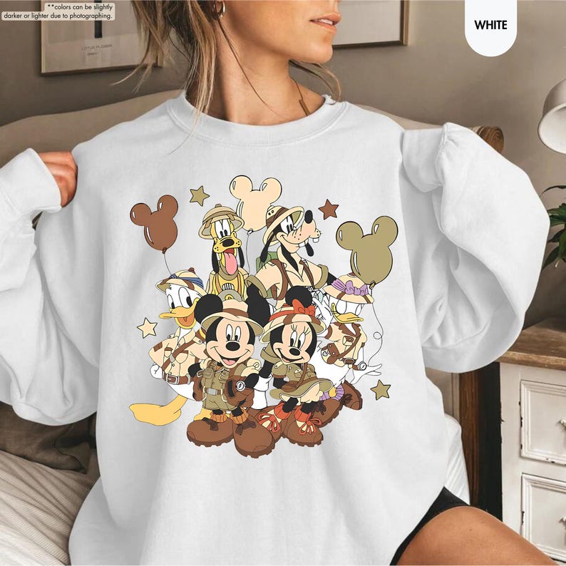 May include: White sweatshirt featuring a colorful graphic of Disney characters in safari outfits. Mickey and Minnie Mouse, Donald Duck, Goofy, and Pluto are depicted with brown safari hats and outfits. Brown Mickey Mouse-shaped balloons and stars complete the design.