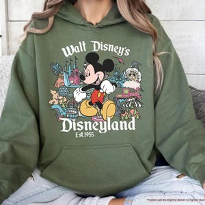 May include: Military green hoodie featuring a colorful graphic of Mickey Mouse in front of Disneyland attractions. The text "Walt Disney's Disneyland Est. 1955" is printed above and below the graphic. The hoodie has a front pocket and a drawstring hood.
