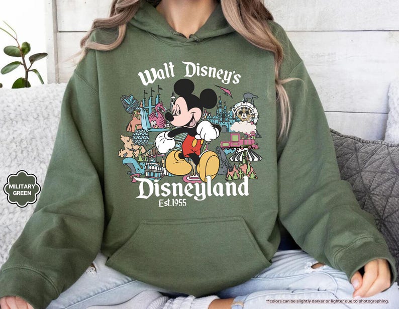 May include: Military green hoodie featuring a colorful graphic of Mickey Mouse in front of Disneyland attractions. The text "Walt Disney's Disneyland Est. 1955" is printed above and below the graphic. The hoodie has a front pocket and a drawstring hood.