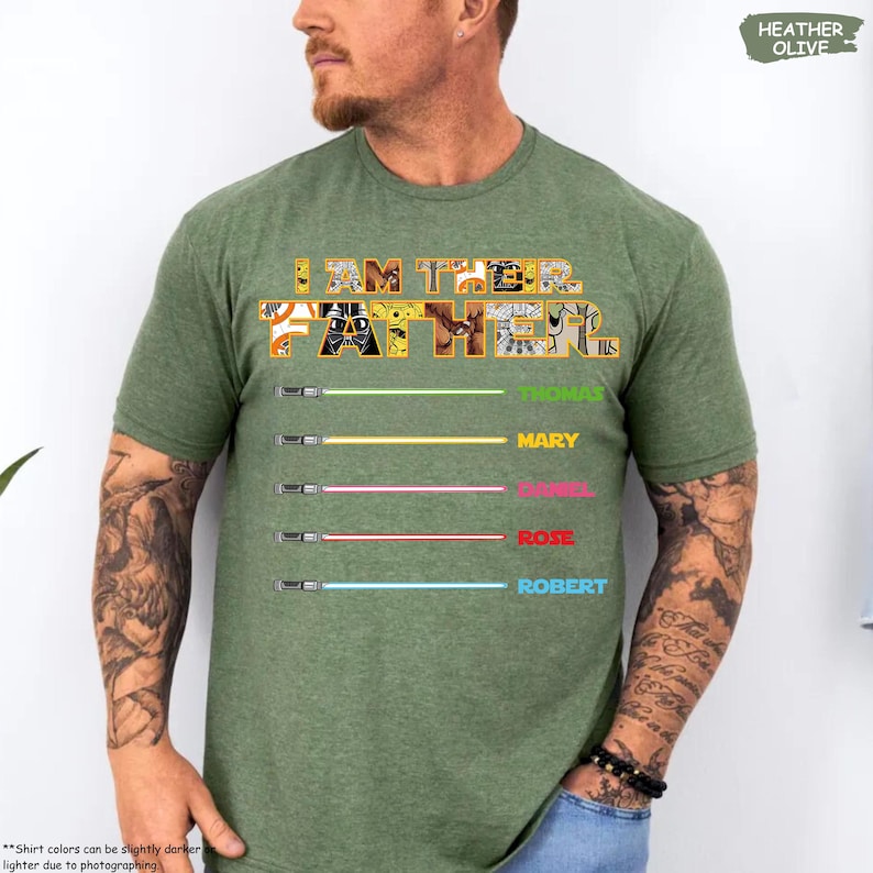 Puede incluir: Camiseta verde oliva jaspeado con el texto "I AM THEIR FATHER" en un dise&ntilde;o multicolor. Debajo, cinco dise&ntilde;os de sables de luz con los nombres: Thomas, Mary, Daniel, Rose y Robert. La camiseta es de material suave.