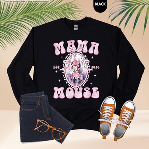 May include: Black sweatshirt with the text "MAMA MOUSE EST 2026" in pink, featuring a Minnie Mouse graphic. The image includes blue jeans, orange eyeglasses, and orange sneakers.