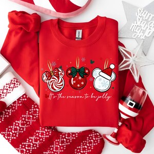 May include: Red sweatshirt with three Mickey Mouse ornament designs. Each ornament has a different design: a candy cane bow, a green bow, and a Santa hat. The text "It's the season to be jolly" is below the ornaments. Christmas theme.