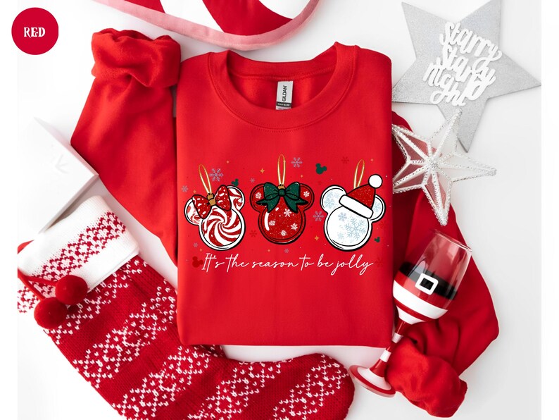 May include: Red sweatshirt with three Mickey Mouse ornament designs. Each ornament has a different design: a candy cane bow, a green bow, and a Santa hat. The text "It's the season to be jolly" is below the ornaments. Christmas theme.