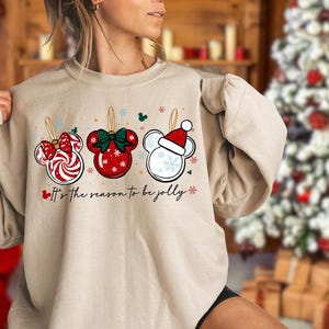 May include: Sand-colored sweatshirt featuring three Mickey Mouse-shaped Christmas ornaments. The ornaments are red, white, and green, with snowflake and bow details. The text "It's the season to be jolly" is printed below the ornaments.