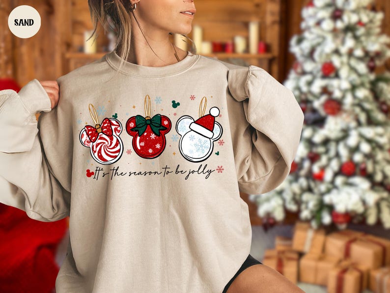May include: Sand-colored sweatshirt featuring three Mickey Mouse-shaped Christmas ornaments. The ornaments are red, white, and green, with snowflake and bow details. The text "It's the season to be jolly" is printed below the ornaments.