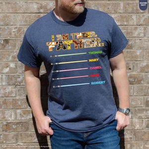 Puede incluir: Camiseta azul marino jaspeado con el texto "I AM THE FATHER" en un dise&ntilde;o colorido tipo collage. Debajo, cinco sables de luz de colores con los nombres: Thomas, Mary, Daniel, Rose y Robert.