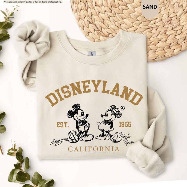 May include: A sand-coloured sweatshirt with the word "DISNEYLAND" in a brown, arched font. Below the text are illustrations of Mickey and Minnie Mouse, with the text "EST. 1955 CALIFORNIA". The sweatshirt has long sleeves and a crew neck.