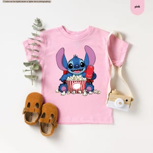 May include: Pink t-shirt featuring a cartoon character holding popcorn and a red soda can. The character is blue with large ears and is sitting in front of a red and white striped popcorn bucket. Brown leather shoes are at the bottom.