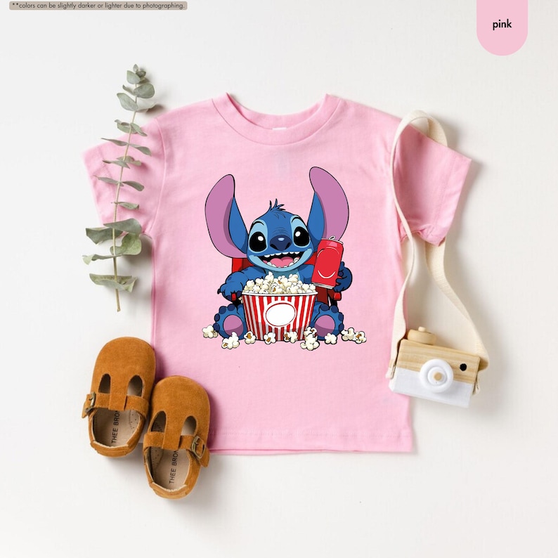May include: Pink t-shirt featuring a cartoon character holding popcorn and a red soda can. The character is blue with large ears and is sitting in front of a red and white striped popcorn bucket. Brown leather shoes are at the bottom.