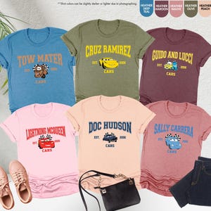 Puede incluir: Varias camisetas con tem&aacute;tica de Cars en varios colores, cada una con el nombre de un personaje y gr&aacute;ficos. Las camisetas incluyen Tow Mater, Cruz Ramirez, Guido y Lucci, Lightning McQueen, Doc Hudson y Sally Carrera. La imagen tambi&eacute;n incluye zapatillas deportivas, un bolso y vaqueros.