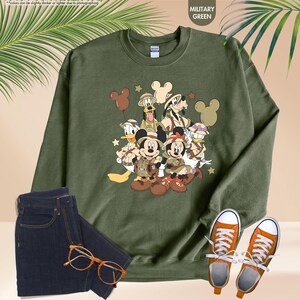 May include: Military green sweatshirt featuring a graphic of Disney characters in safari outfits. The design includes Mickey and Minnie Mouse, Donald Duck, Goofy, and Pluto. The sweatshirt is paired with dark blue jeans and orange sneakers.