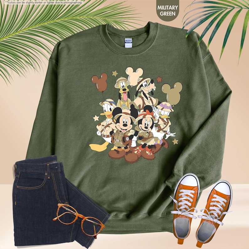 May include: Military green sweatshirt featuring a graphic of Disney characters in safari outfits. The design includes Mickey and Minnie Mouse, Donald Duck, Goofy, and Pluto. The sweatshirt is paired with dark blue jeans and orange sneakers.