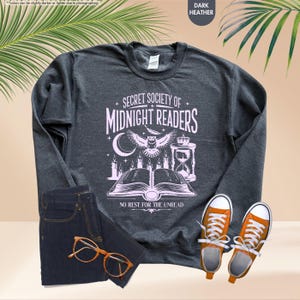 May include: Dark heather gray sweatshirt with the text "SECRET SOCIETY OF MIDNIGHT READERS" in white. The graphic includes an owl, candles, an hourglass, and an open book. The sweatshirt is paired with dark blue jeans and orange sneakers.