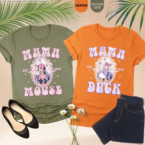 May include: Two graphic t-shirts, one olive green and one orange, with the text "MAMA" and character graphics. The olive shirt says "MOUSE" and the orange shirt says "DUCK". Both shirts have "EST 2026" printed on them. Black flats and denim jeans are also in the image.
