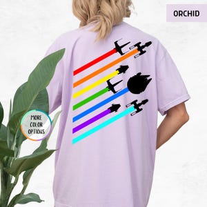 K&ouml;nnte beinhalten: Ein lila T-Shirt mit einem Regenbogenmuster und Silhouetten von Raumschiffen. Der Text "ORCHID" befindet sich in der oberen rechten Ecke des Bildes.