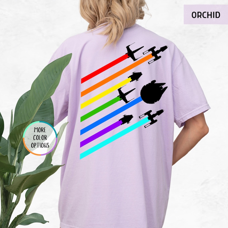 K&ouml;nnte beinhalten: Ein lila T-Shirt mit einem Regenbogenmuster und Silhouetten von Raumschiffen. Der Text "ORCHID" befindet sich in der oberen rechten Ecke des Bildes.