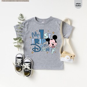 K&ouml;nnte beinhalten: Graues T-Shirt mit kurzen &Auml;rmeln und einem blauen und wei&szlig;en Aufdruck mit der Aufschrift "My 1st Trip to Disney" und einem Comic-Mickey-Mouse-Kopf.