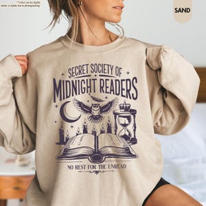 May include: Sand-colored sweatshirt with a graphic design featuring the text "SECRET SOCIETY OF MIDNIGHT READERS." The design includes an owl, open book, candles, and an hourglass. The phrase "NO REST FOR THE UNREAD" is at the bottom.