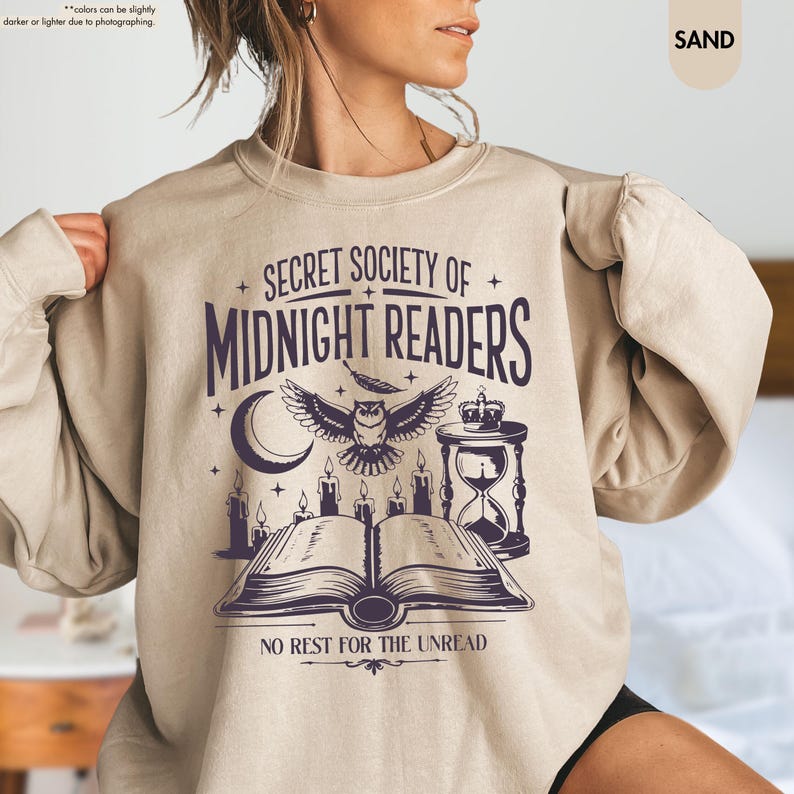 May include: Sand-colored sweatshirt with a graphic design featuring the text "SECRET SOCIETY OF MIDNIGHT READERS." The design includes an owl, open book, candles, and an hourglass. The phrase "NO REST FOR THE UNREAD" is at the bottom.