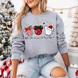 May include: A gray sweatshirt featuring three Mickey Mouse-shaped Christmas ornaments. The ornaments are red, white, and green, with a candy cane, bow, and Santa hat design. The text "It's the season to be jolly" is printed below the ornaments.